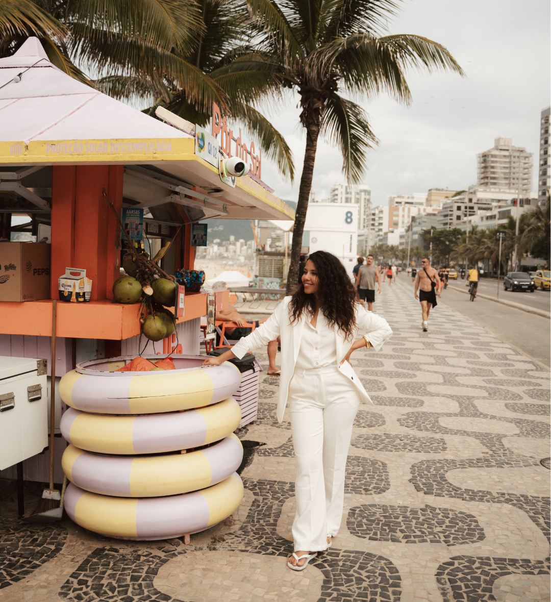 A woman in a white two-piece outfit stands on a beach boardwalk beside a small food stall with stacked yellow and white float rings, palm trees, and distant city buildings.