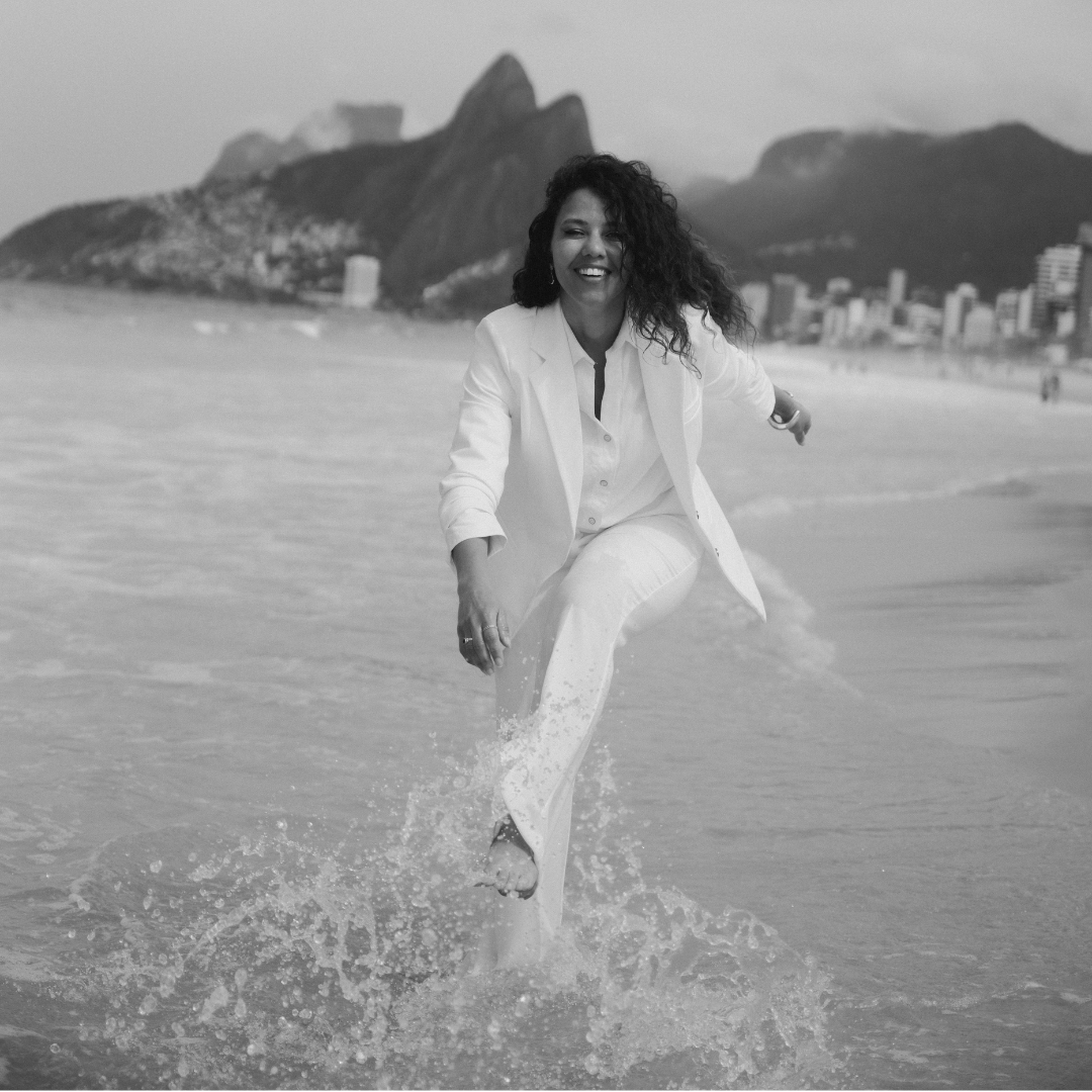 A black and white lifestyle photograph of a woman in a white suit running through splashing ocean water on a beach, with mountains and city buildings in the background.