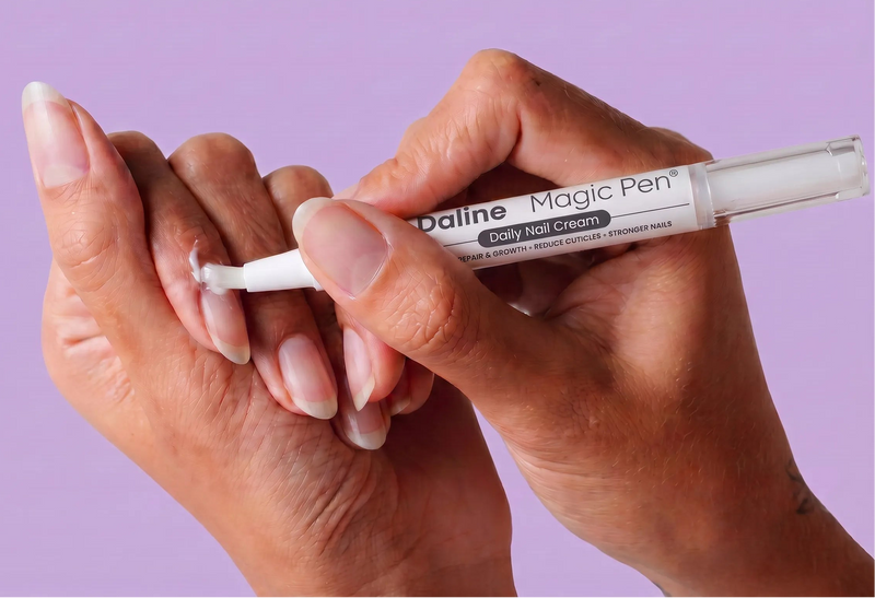 A close-up of a person's hands applying a white nail cream from a Daline Magic Pen to a fingernail, set against a solid lavender background.
